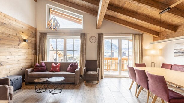 A spacious living area with a wooden ceiling, a sofa, a chair, a dining table with chairs, and large windows offering a view of mountains. - Superior Ferienhaus # 4C mit Sauna (Uttendorf)