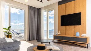 A modern living room with a sofa, two round tables, a TV mounted on a wooden panel, and a view of the sea through large windows. - Luxury Villa with pool and panoramic sea view! (Potomje)