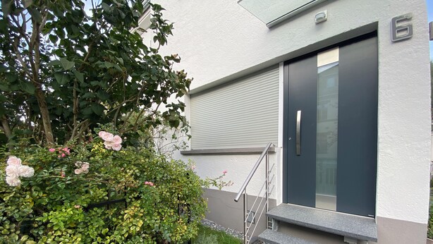 Een modern gebouw met een grijze gevel, een zwarte deur en een kleine trap van natuursteen die naar de ingang leidt.