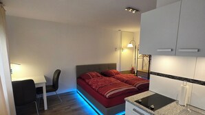 Een kleine hotelkamer met een bed, bureau, stoel, kitchenette en badkamer.