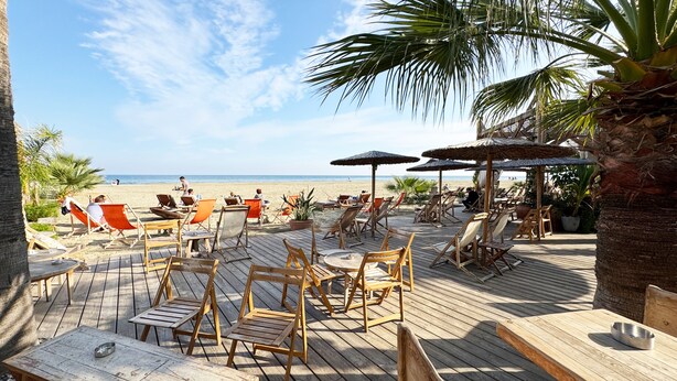Une terrasse en bois avec des chaises longues et des tables, donnant sur une plage parsemée de parasols et de palmiers.