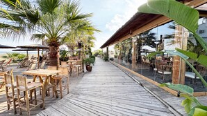 Une terrasse en bois, agrémentée de tables et de chaises, entourée de palmiers et de parasols, avec vue sur une plage.