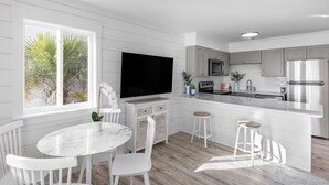 Una cucina moderna con mobili bianchi, una grande isola con sgabelli, una TV a schermo piatto e vista su palme attraverso la finestra.