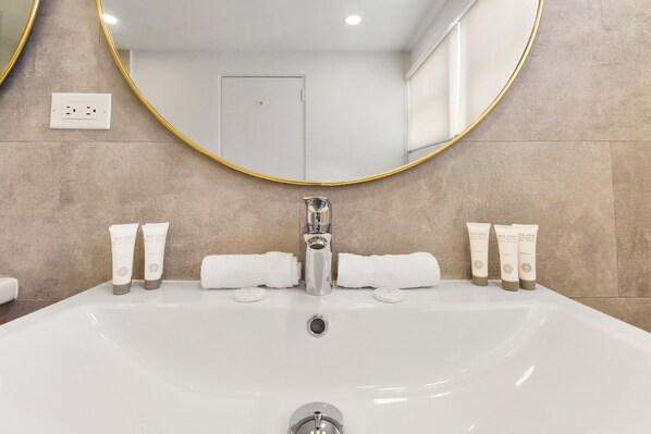 Une salle de bain avec un grand lavabo blanc, un miroir rond doté d’un cadre doré et des articles de toilette posés sur le comptoir.