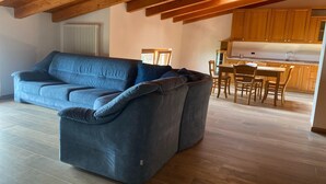 Een moderne woonkamer met een grote grijze bank, houten balken in het plafond, een eethoek met houten stoelen en een tafel, en een keuken met houten kastjes.