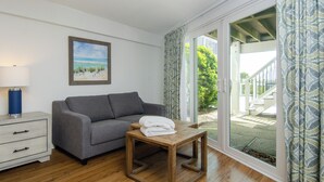 Ein Zimmer mit grauem Sofa, einem Holztisch mit Handtüchern, einem weißen Nachttisch, einer Lampe und einem großen Fenster mit gemusterten Vorhängen.