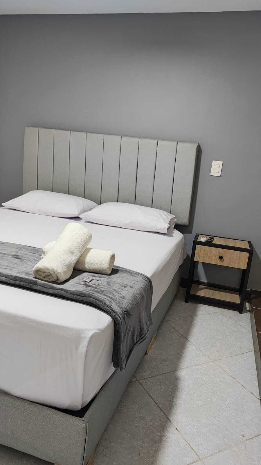 A bed with a grey headboard, white bedding, and two rolled towels. A small wooden nightstand with a drawer is beside the bed.