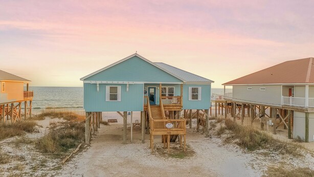 Ein Strandhaus mit Holzterrasse und Veranda, auf Stelzen über dem Sand erhöht.
