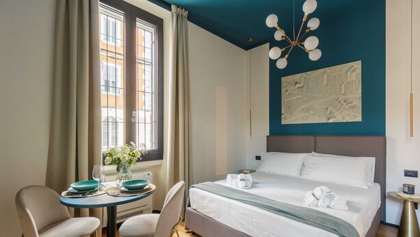A hotel room with a bed, a dining table with chairs, a window with curtains, and a modern chandelier. - Bright Ground-floor Studio in Milan (Milan)