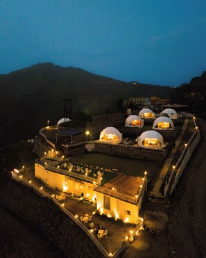 A series of dome-shaped accommodations with outdoor seating areas, surrounded by a stone wall and set against a mountainous backdrop. - Amantra Hills Rishikesh (Narendranagar)