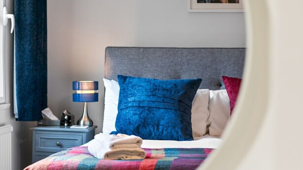 A neatly made bed with a colorful quilt, a blue pillow, and a white pillow. There is a nightstand with a lamp and a phone.