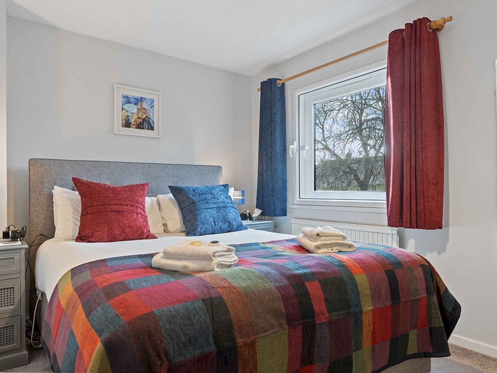 Een net opgemaakt bed met een kleurrijk geruit plaid, een hoofdbord en kussens in rood en blauw. Er is een raam met uitzicht op bomen en een rood gordijn.