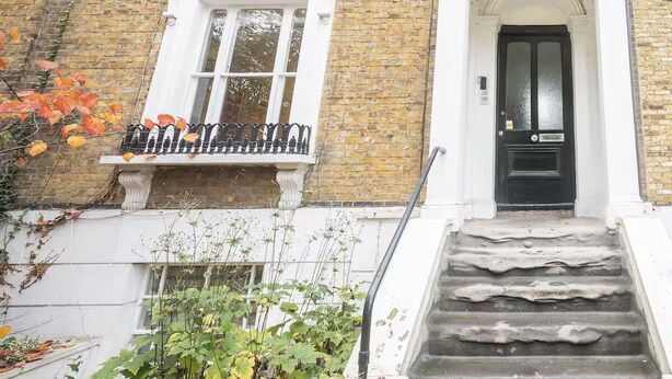 A traditional brick building with a white window and black door, a small balcony with a metal railing, and a set of stone steps leading to the entrance. - Brockley Victorian apartment train links London - Pass the Keys (London)