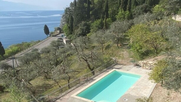 Una piscina rectangular rodeada por una terraza de piedra, con vista al mar y a una colina con árboles.