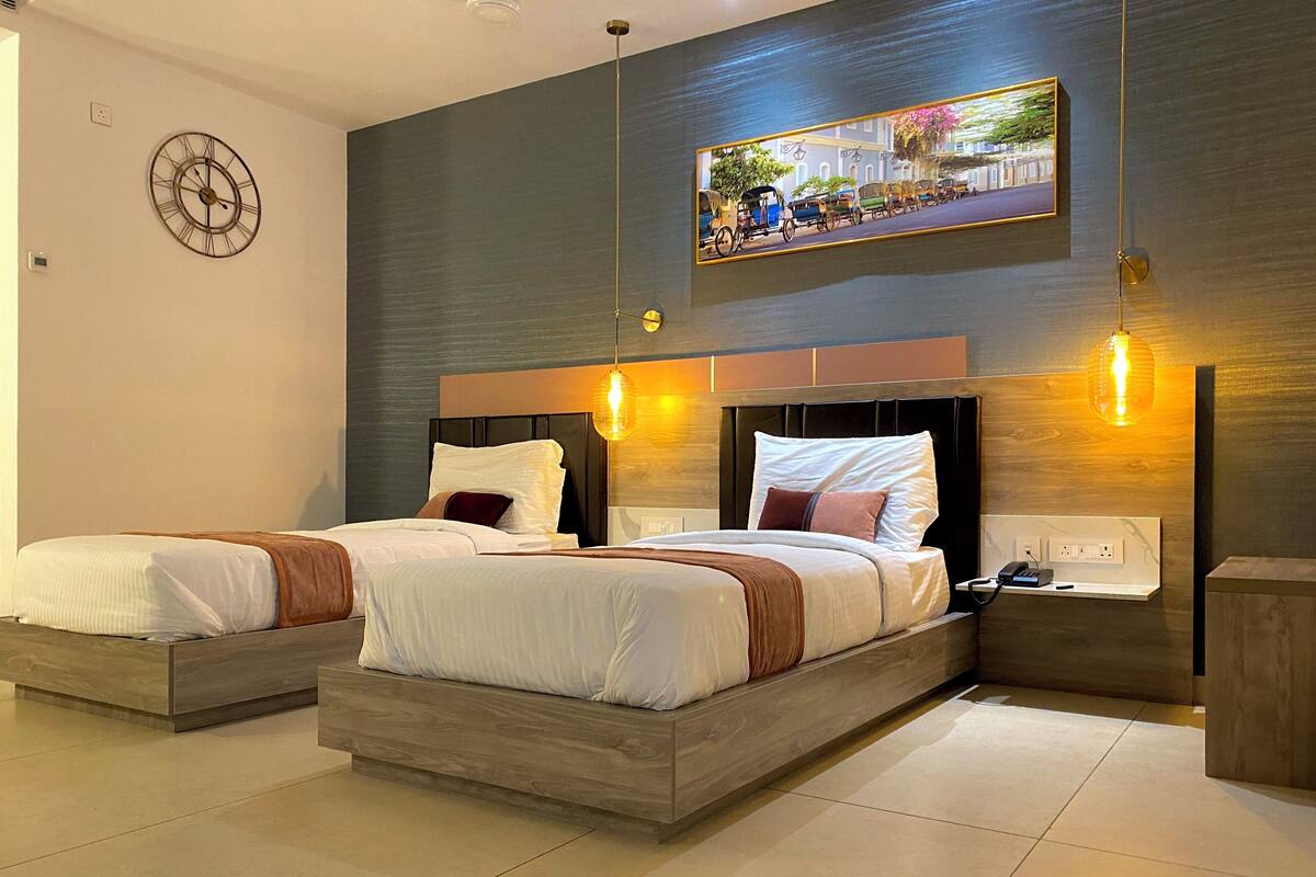 A modern hotel room with two beds, a large wall clock, a framed cityscape picture, and a nightstand with a phone and charger.