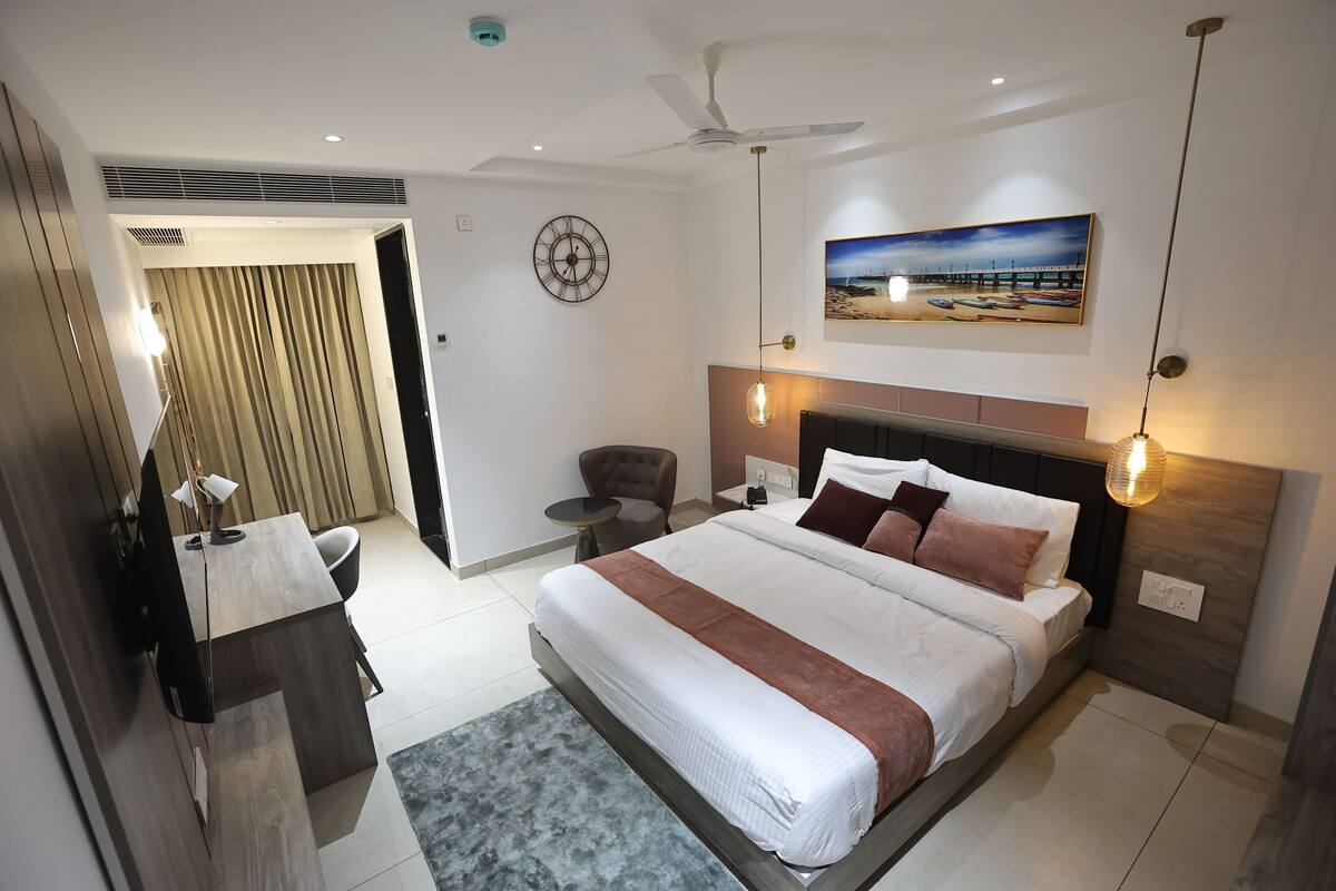 A modern hotel room with a large bed, a desk, a chair, a ceiling fan, a painting, and a clock on the wall.