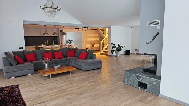 Ein modernes Wohnzimmer mit einem grauen Ecksofa, einem Holztisch und einem Kamin. Eine Treppe führt zu einer oberen Ebene, und im Hintergrund ist eine Küche zu sehen.