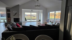 Ein geräumiges Wohnzimmer mit einem großen, schwarzen Ecksofa, einem Kamin und Blick auf ein Feld durch große Fenster.