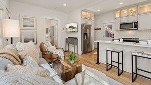 A modern kitchen with white cabinets, a stainless steel refrigerator, and a microwave. There is a wooden coffee table with a plant, and a bar stool. - Chic Luxury Retreat – Old Town Carriage House (Fort Collins)