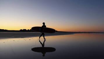 Eine Person geht bei Sonnenuntergang am Strand mit einem Surfbrett.