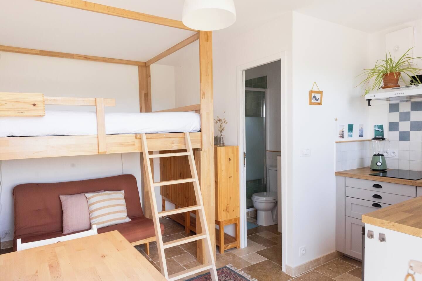Una habitación compacta con una cama nido, una escalera de madera, un sofá, una cocina con un lavabo y una licuadora, y un baño con inodoro.