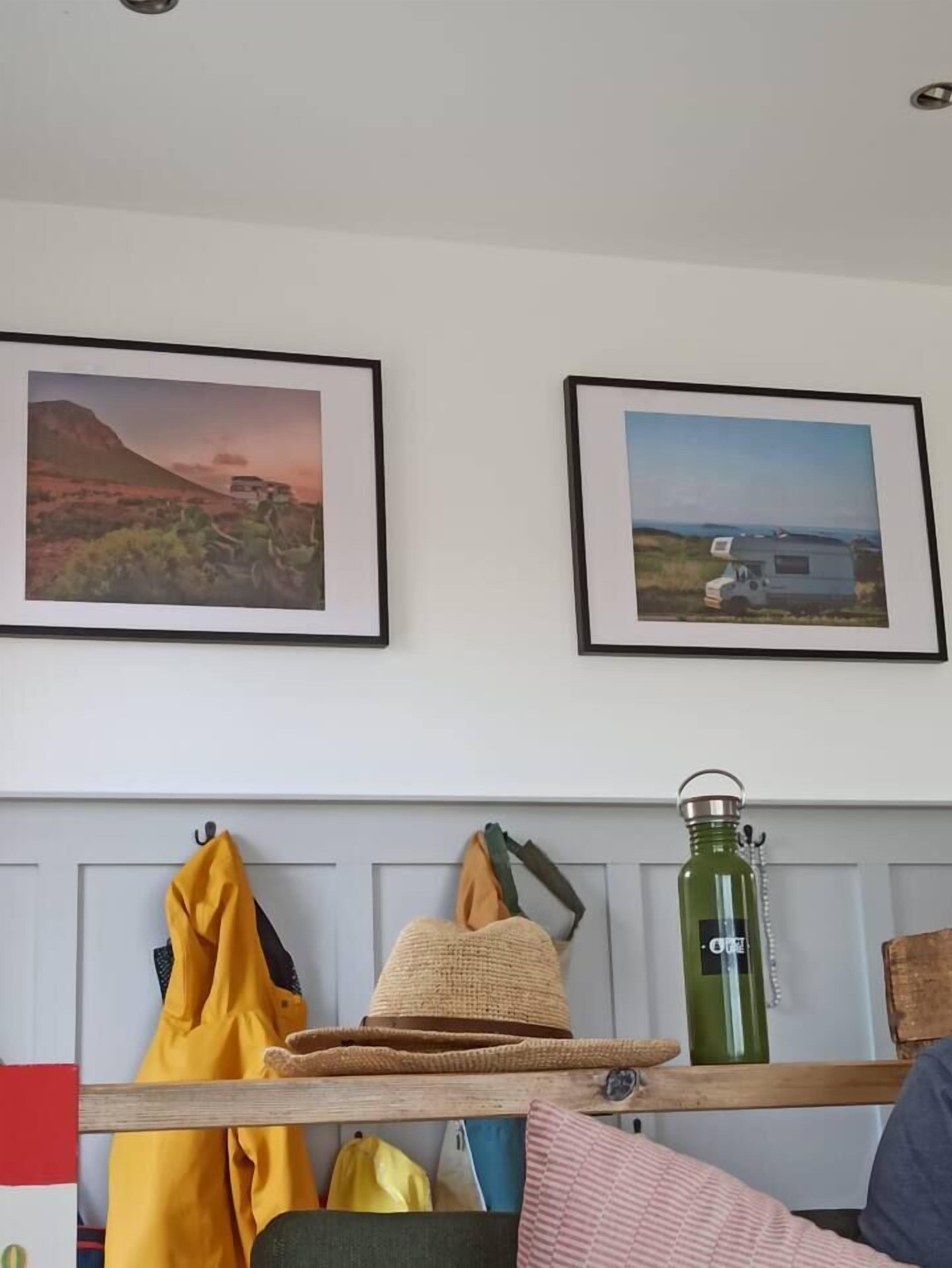 A room with two framed pictures on the wall, a wooden bench with a hat and a jacket, and a green water bottle.