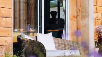 Ein Korbsessel mit Kissen auf einer Holzterrasse, eine Glastür zur Inneneinrichtung und eine Topfpflanze im Vordergrund.