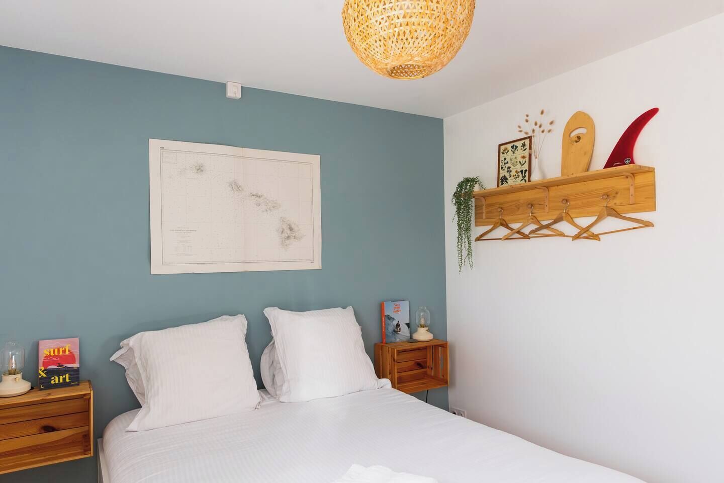 A bedroom with a bed, a map on the wall, a wooden shelf with surfboards, and a hanging plant.