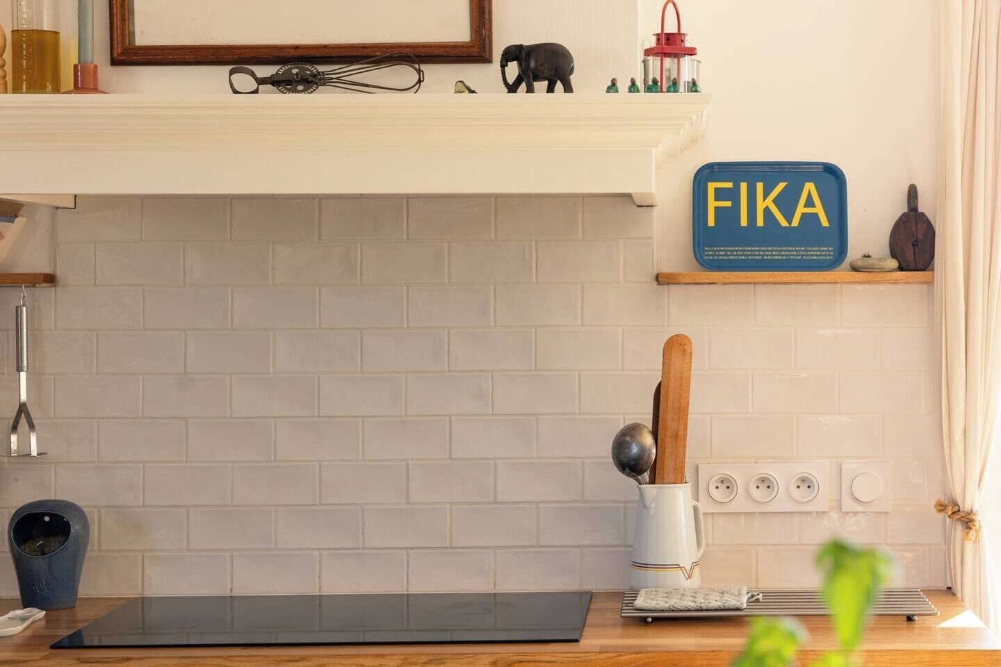 Cozinha com painel de tijolos brancos, bancada preta, placa FIKA e uma colher de pau em caneca branca.