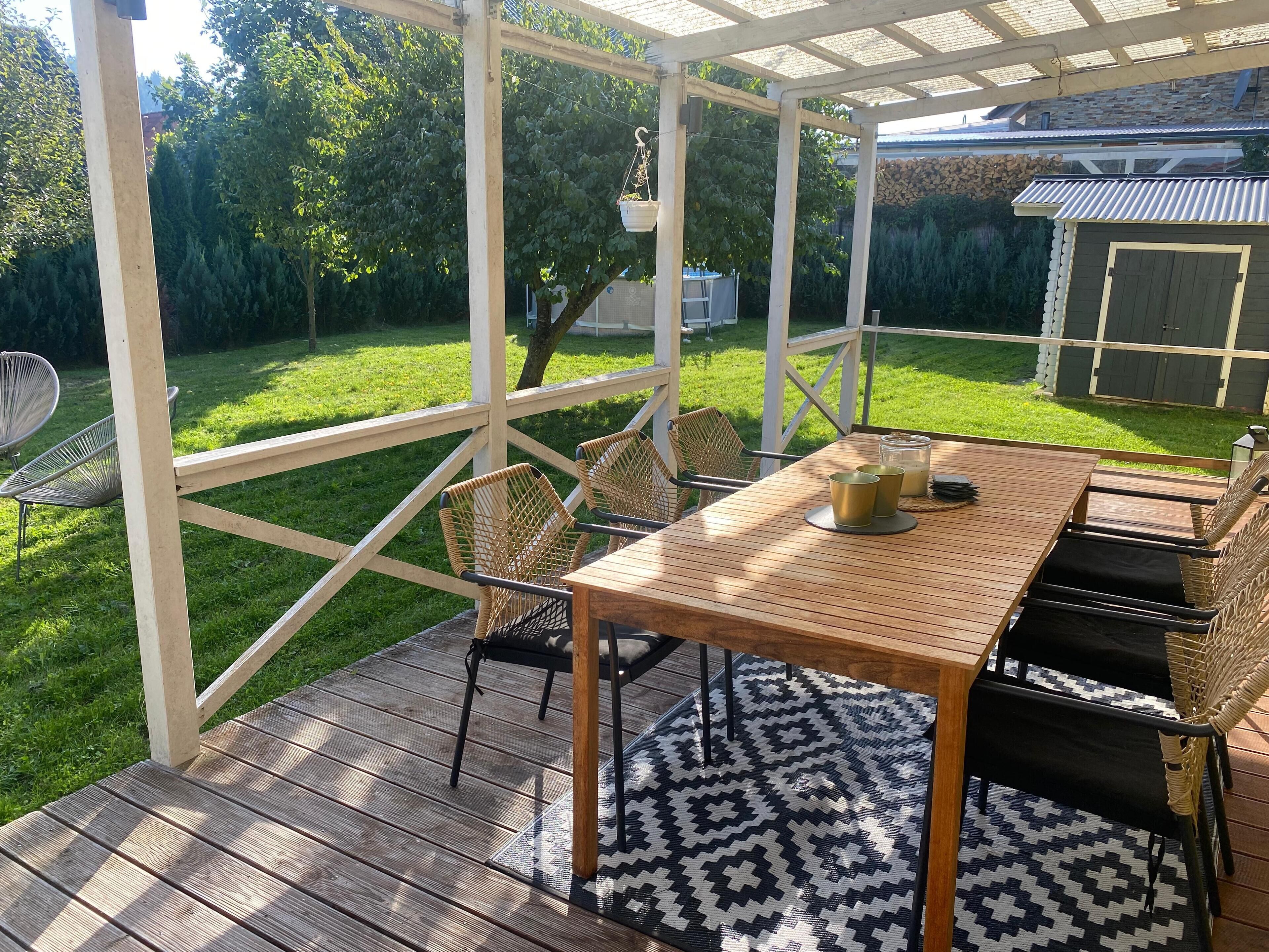 En täckt uteplats med ett träbord och stolar, en mönstrad matta och utsikt över en grön gräsmatta och träd.