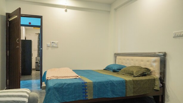 A bedroom with a bed, a chair, and a door leading to another room. - VJ Rooms (Bengaluru)
