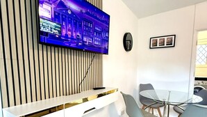 A modern living room with a flat-screen TV mounted on a wooden panel wall, a white entertainment unit, a glass-top dining table with four chairs, and a bench. - Spacious 2 Bedroom flat with Balcony – 5 min to Kennington Tube, Central London (London)