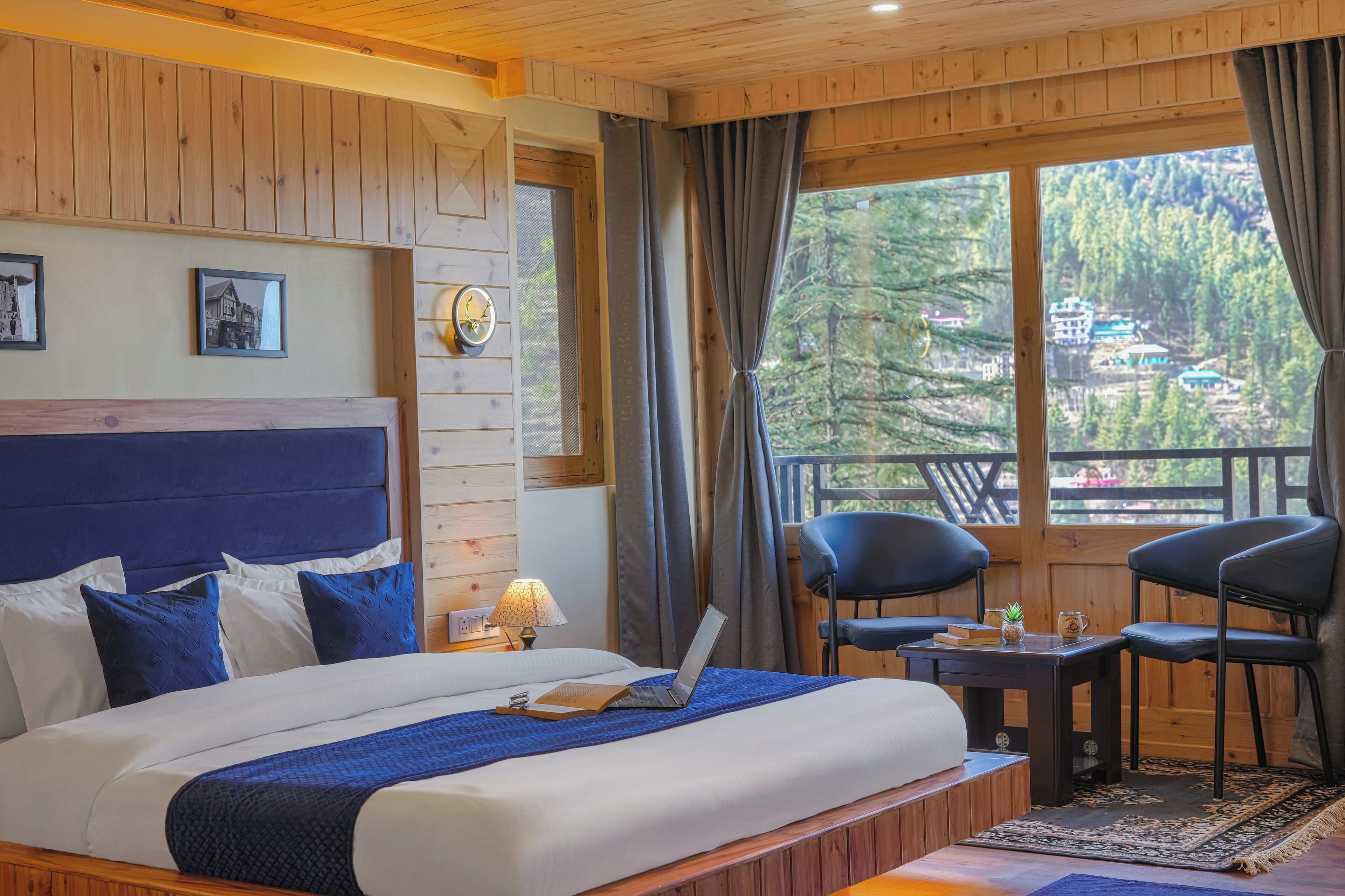 A bedroom with a large bed, a desk, two chairs, a laptop, and a view of a forested area.