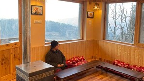 A cozy cabin interior with a wood stove, wooden benches, and large windows offering mountain views.