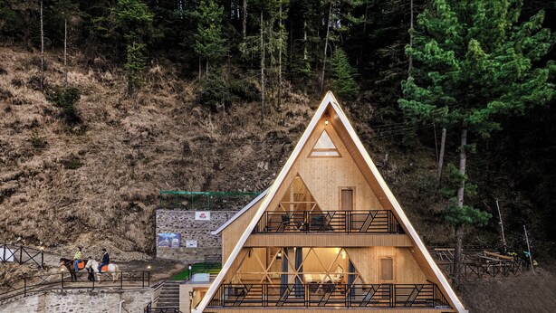 A wooden cabin with a balcony, situated on a hillside with a forest backdrop.