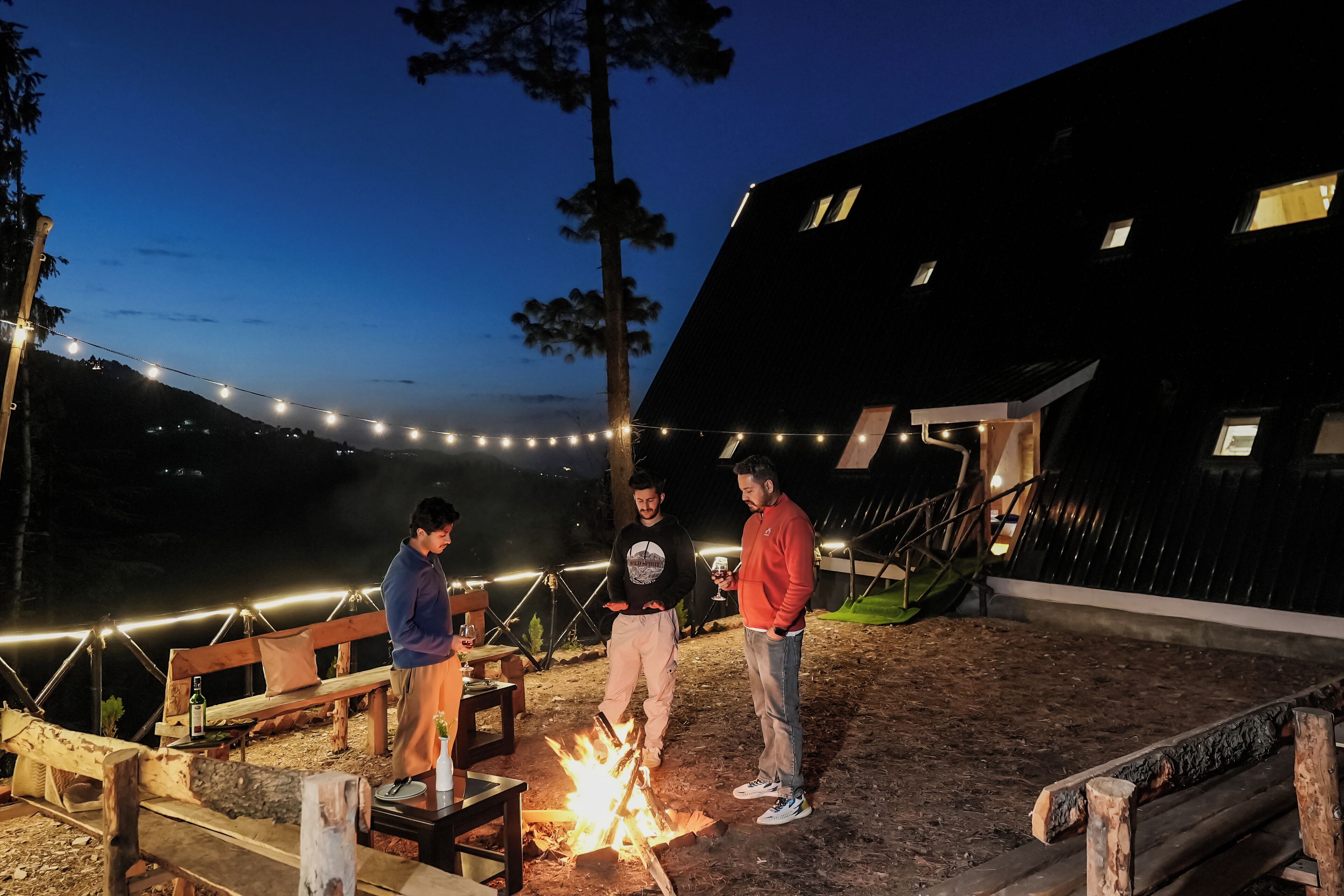 A group of people around a campfire in an outdoor setting with string lights and wooden benches.