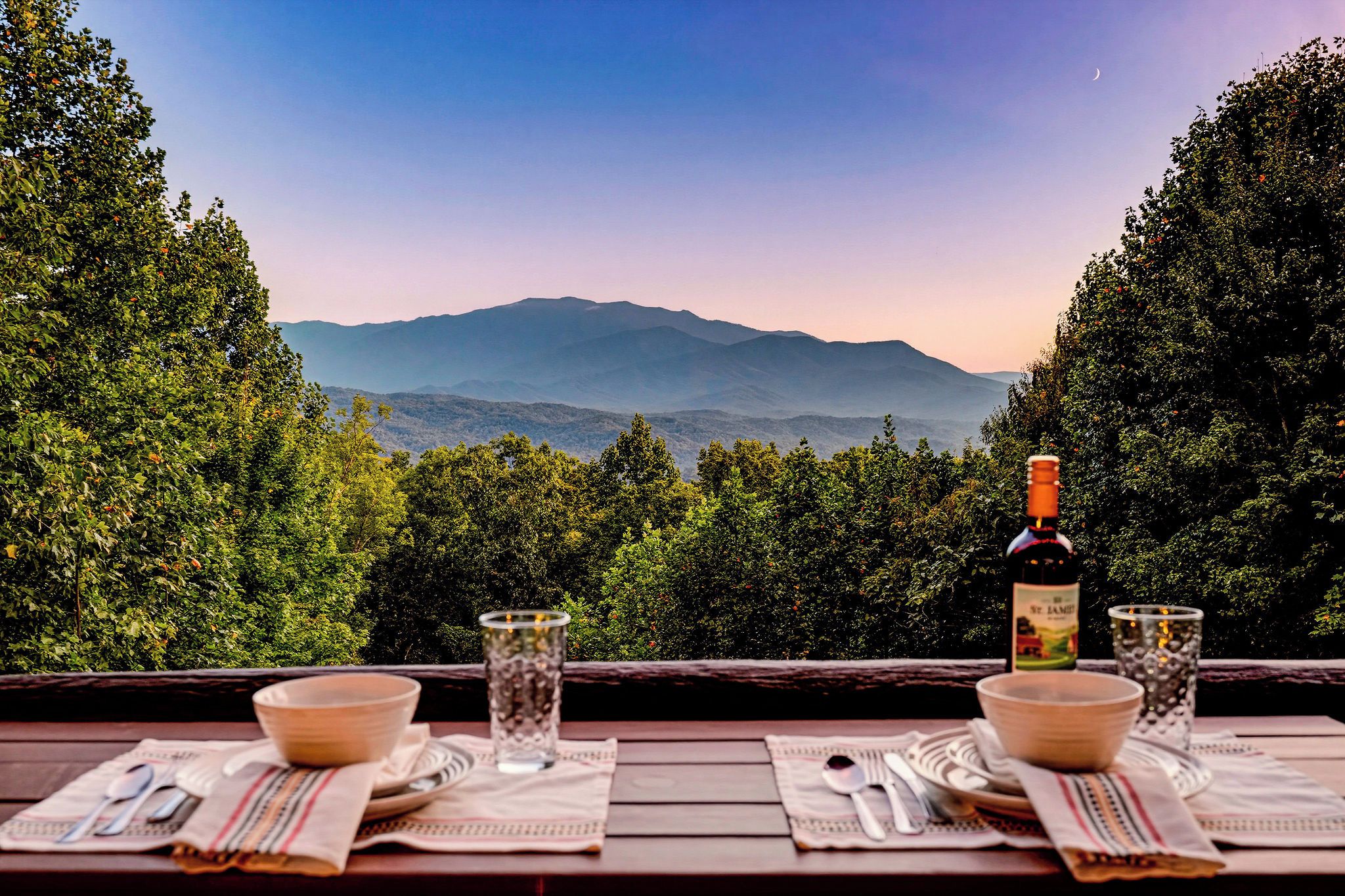 A table set with bowls, glasses, and a bottle, overlooking a mountainous landscape.