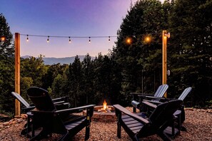 An outdoor seating area with Adirondack chairs around a fire pit, surrounded by trees and a mountain view.