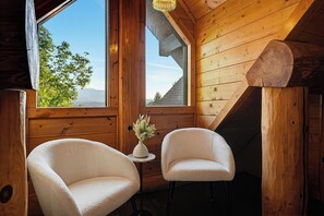 A cozy cabin interior with two plush armchairs, a small table with a vase, and a large window offering a view of the outdoors.