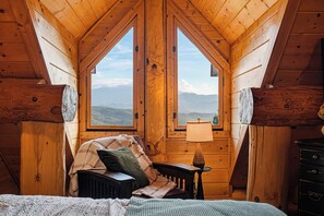 A cozy attic bedroom with a bed, a chair, a table, and a lamp. There are two windows providing a view of the mountains.