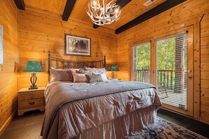 A wooden cabin bedroom with a bed, two bedside lamps, a framed picture on the wall, and a large window with blinds. - Round Log Cabin with Stunning Views (Gatlinburg)