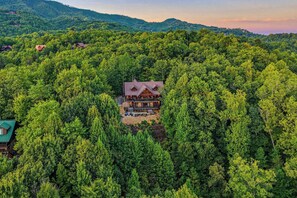 Ein großes, zweistöckiges Holzhaus, umgeben von dichtem Wald, mit einer breiten Veranda und zahlreichen Fenstern.