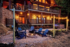 A two-story log cabin with a stone exterior, outdoor seating area with Adirondack chairs, and a fire pit.