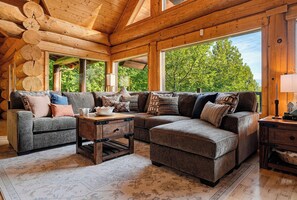 Een blokhut interieur met een hoekbank, een bijzettafeltje en een groot raam met uitzicht op bomen.