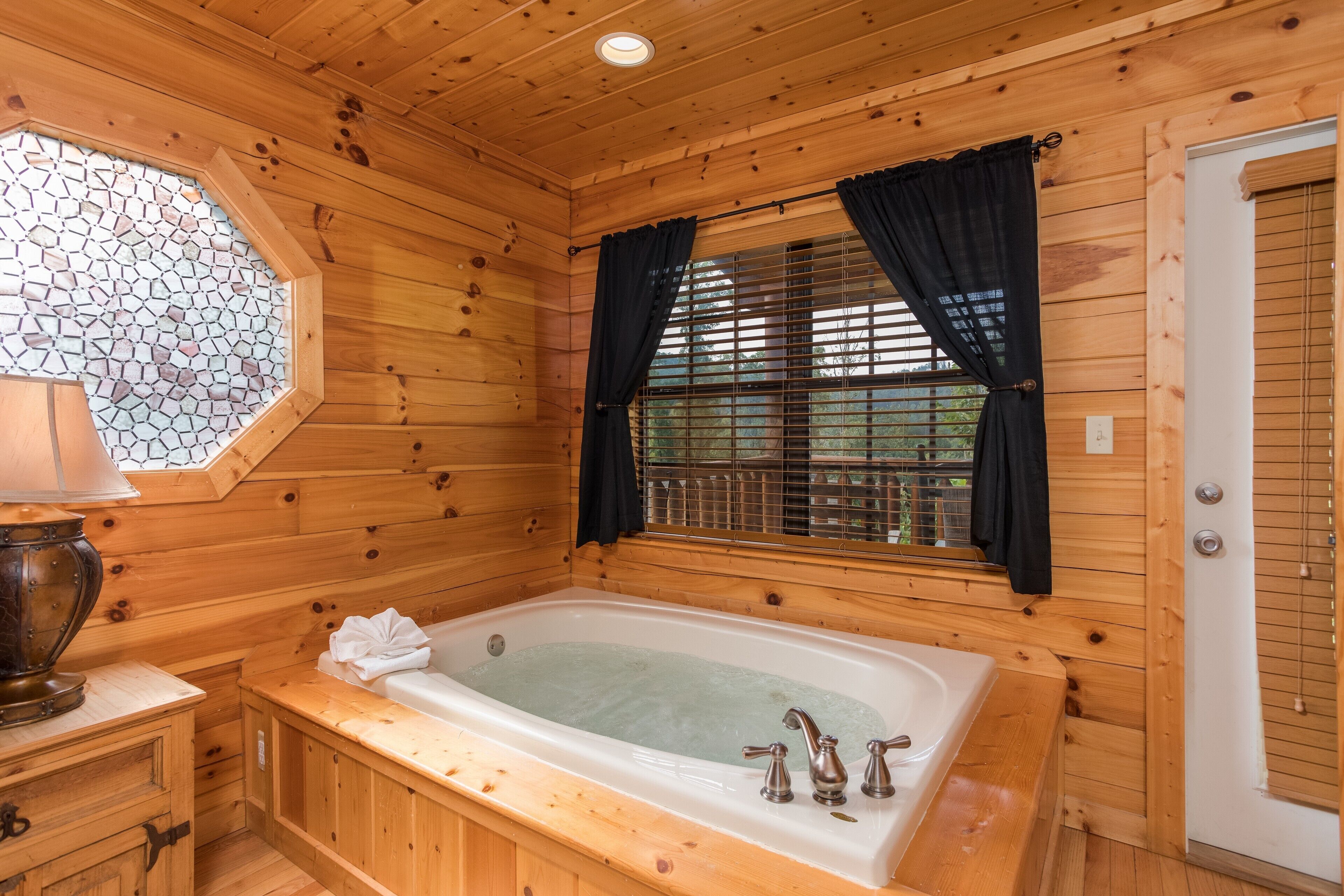 Un baño de cabaña de madera con una bañera, un ventanal con cortinas y un tragaluz con mosaicos.