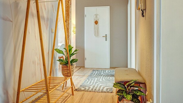 Un couloir avec un fauteuil suspendu en bois, une plante en pot, un tapis à motifs et une porte blanche ornée d’un élément décoratif en macramé.