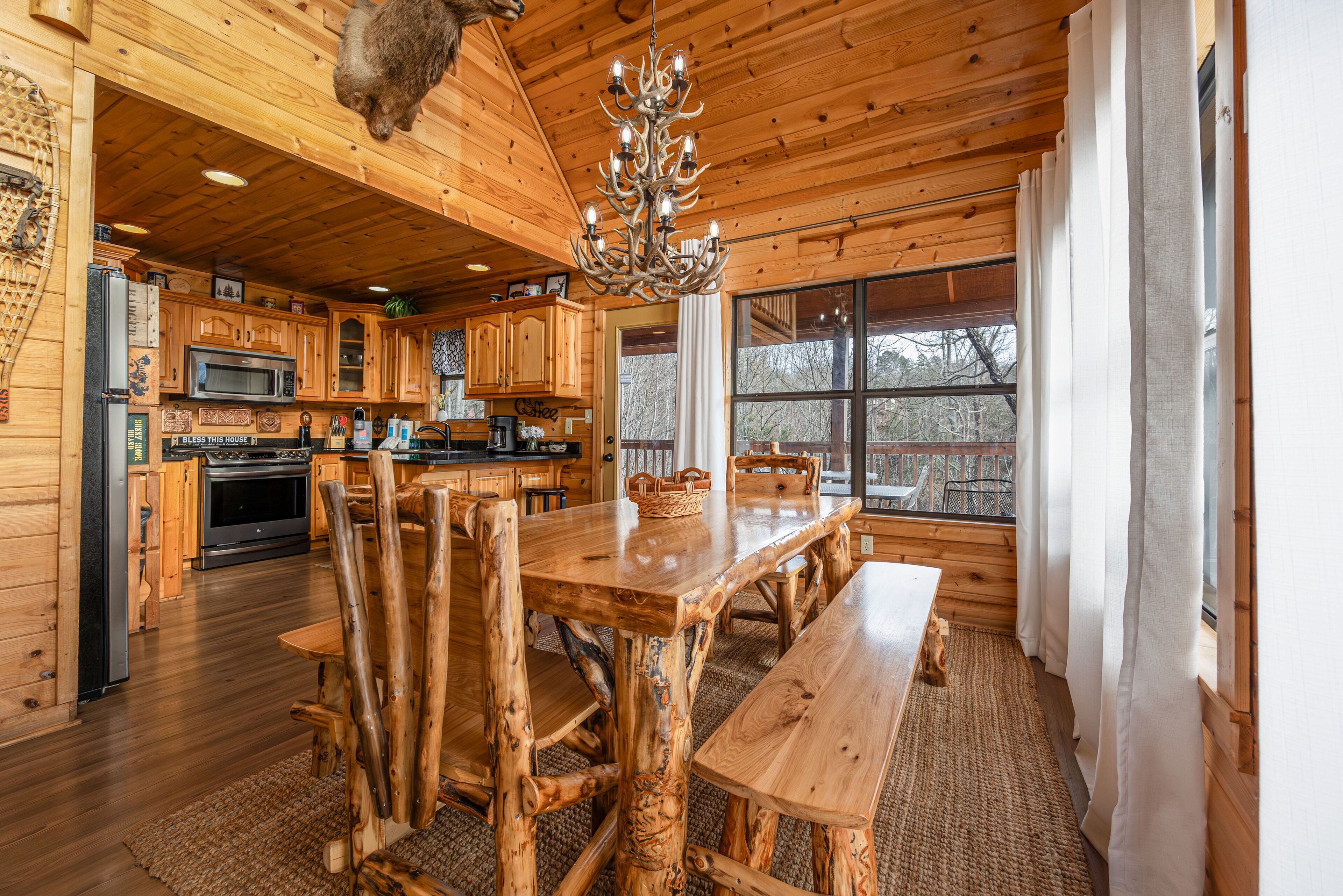 A rustic wooden interior with a large dining table, wooden chairs, a chandelier, and a kitchen area with wooden cabinets and appliances.