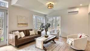 A spacious living room with a brown sofa, a white armchair, and a coffee table. There are large windows providing natural light and a view of trees outside. - 1 month for the price of 1 week! (Mont-Tremblant)