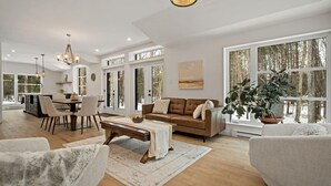 A spacious living room with a large window, a brown leather sofa, a dining table with chairs, and a chandelier. - 1 month for the price of 1 week! (Mont-Tremblant)