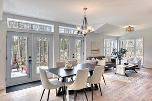 A spacious living room with a large dining table, comfortable seating, and large windows offering a view of the outdoors. - 1 month for the price of 1 week! (Mont-Tremblant)